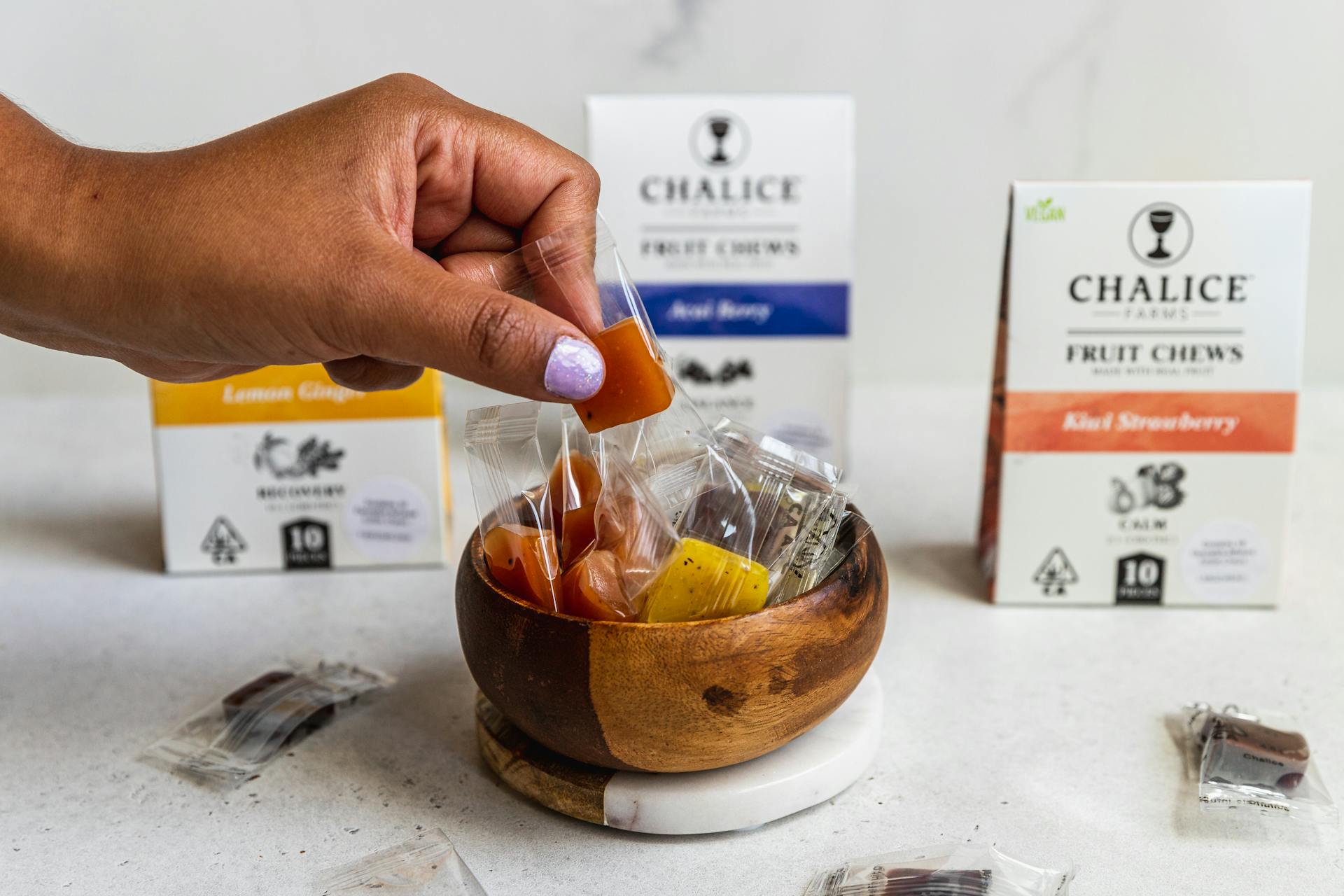 A hand picks up fruit chew candies from a wooden bowl, with packaging visible in the background.