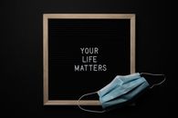Overhead arrangement of framed black photo with white words Your Life Matters on black background near medical mask