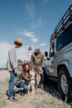 Friends and a dog relax beside a car near a lighthouse on a sunny beach day.