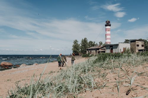 People Walking on Beach Shore Near Lighthouse Under Blue Sky