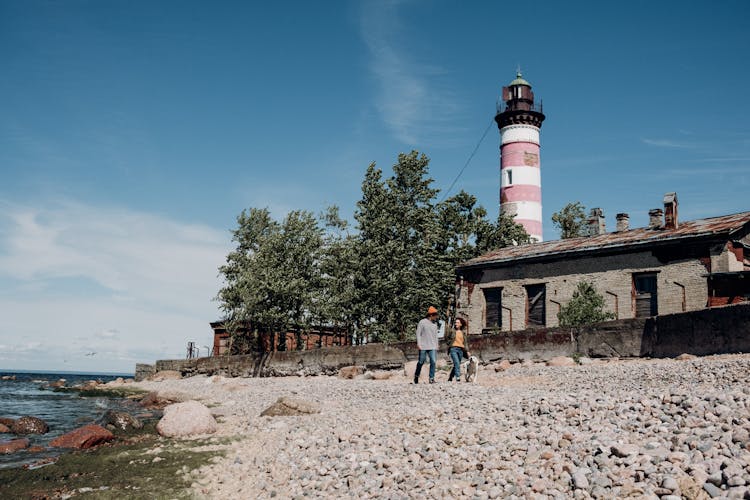 Man And Woman Walking On Shore With A Dog Near Lighthouse