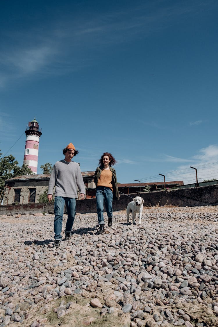 Man And Woman Walking On Rocky Shore With A Dog