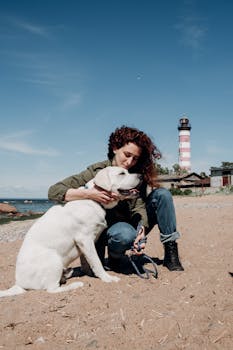 在一个阳光明媚的日子里，一名妇女在海滩上拥抱她的拉布拉多犬，灯塔作为背景