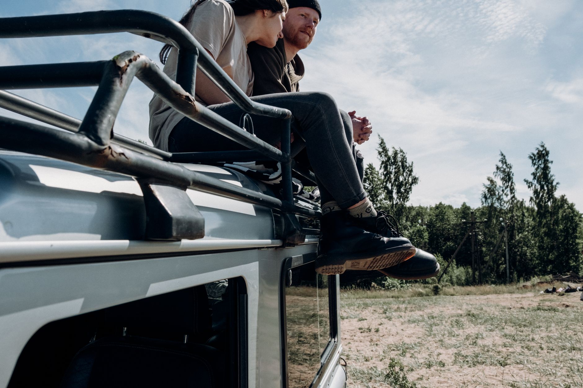 A couple sits on a car roof, enjoying a serene outdoor setting with trees and clear skies.
