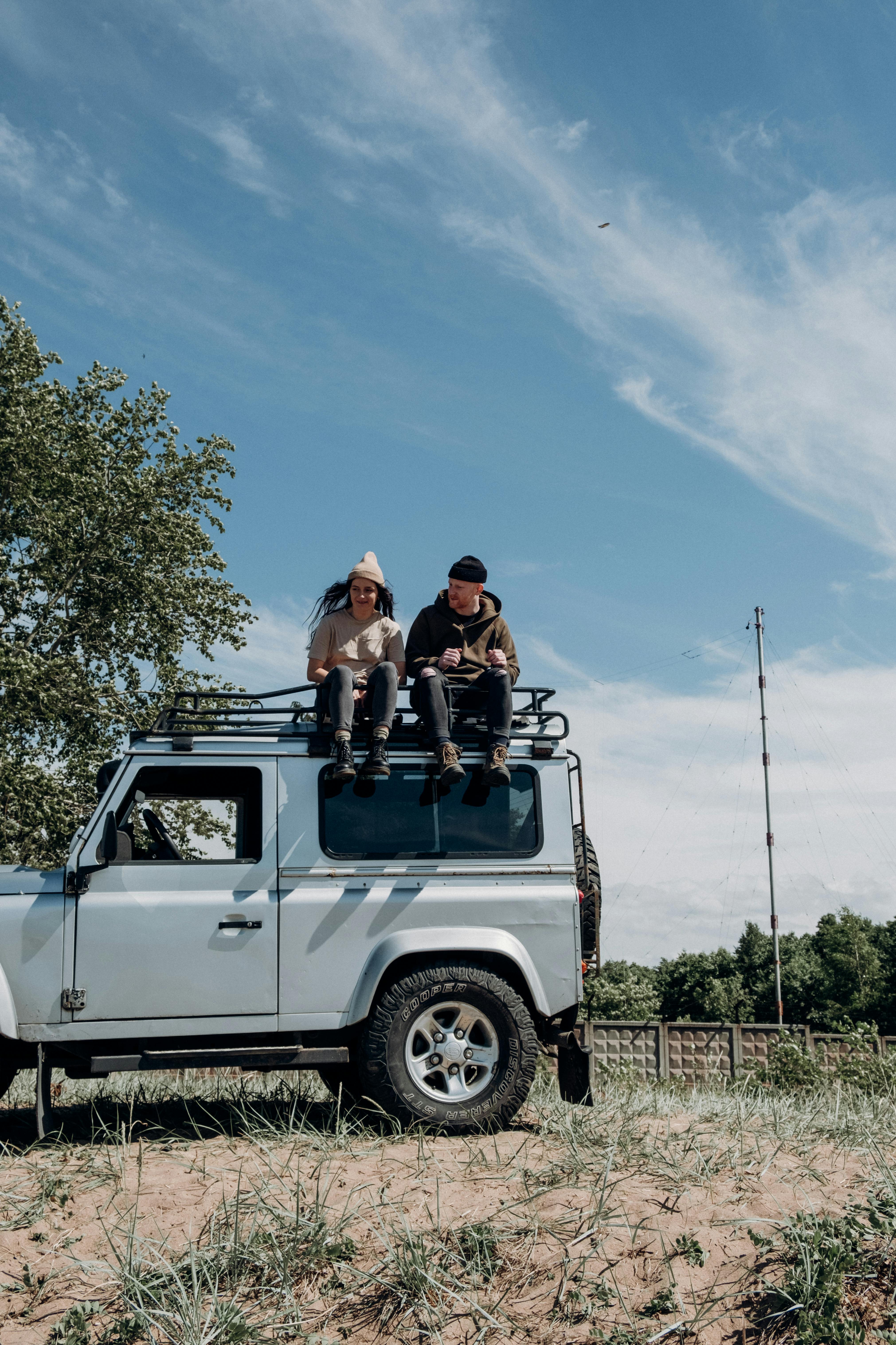 Two young adults sitting on an SUV, enjoying a sunny outdoor road trip.