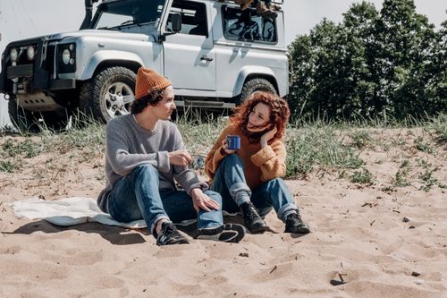 Free Young couple sitting on a sandy beach, enjoying coffee with a jeep in the background. Stock Photo