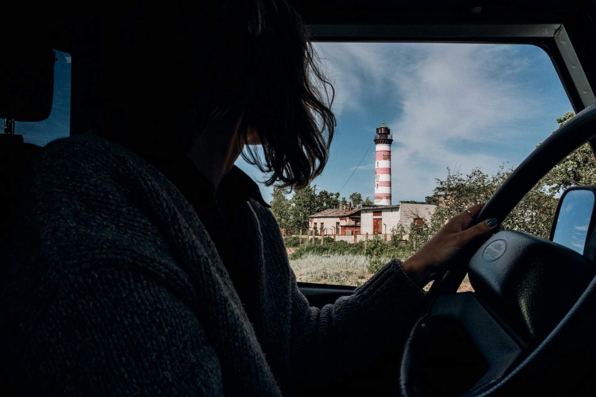 A woman driving with a view of a striped lighthouse through the car window.