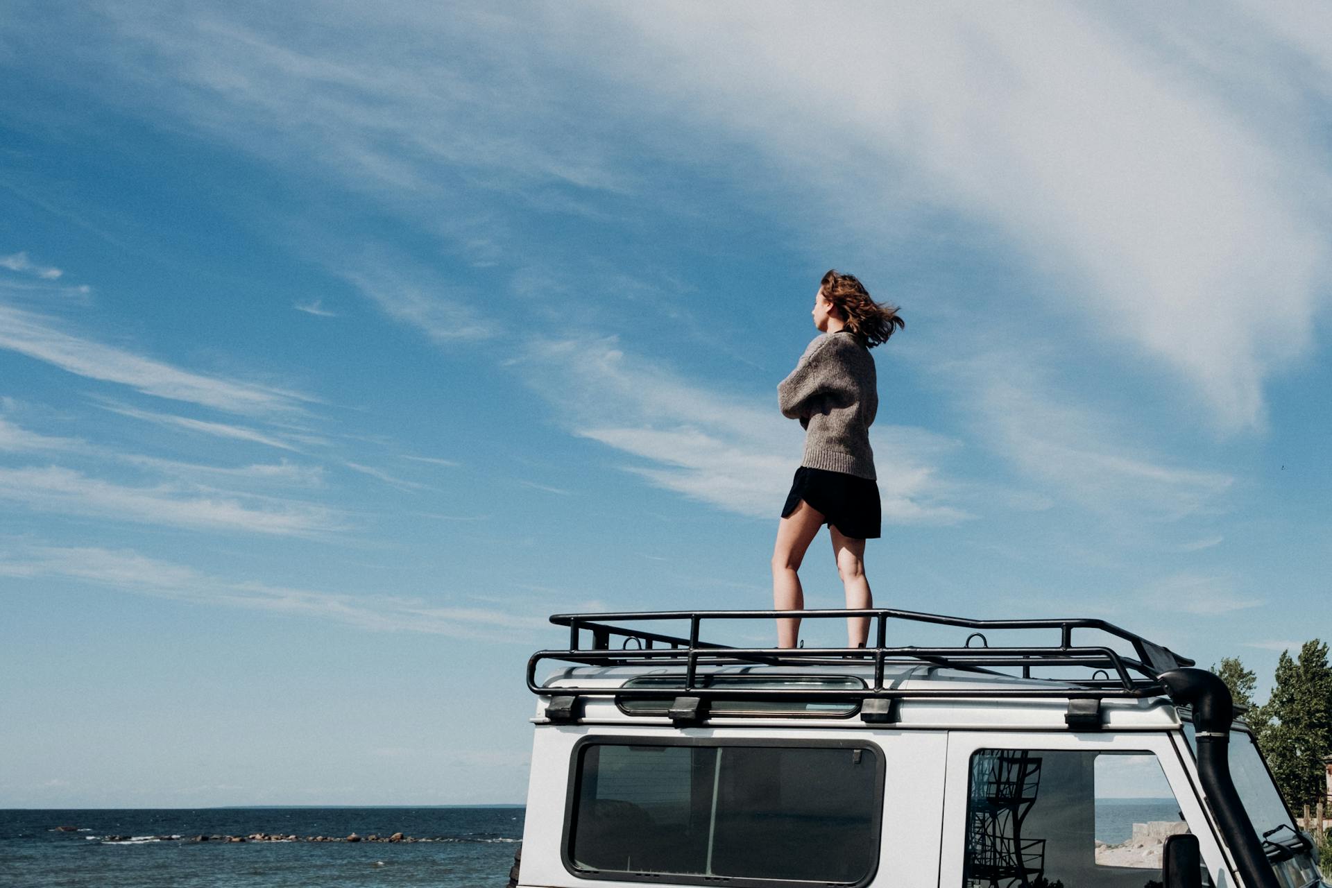 A woman stands confidently on a car roof by the sea, enjoying the freedom under a blue sky.