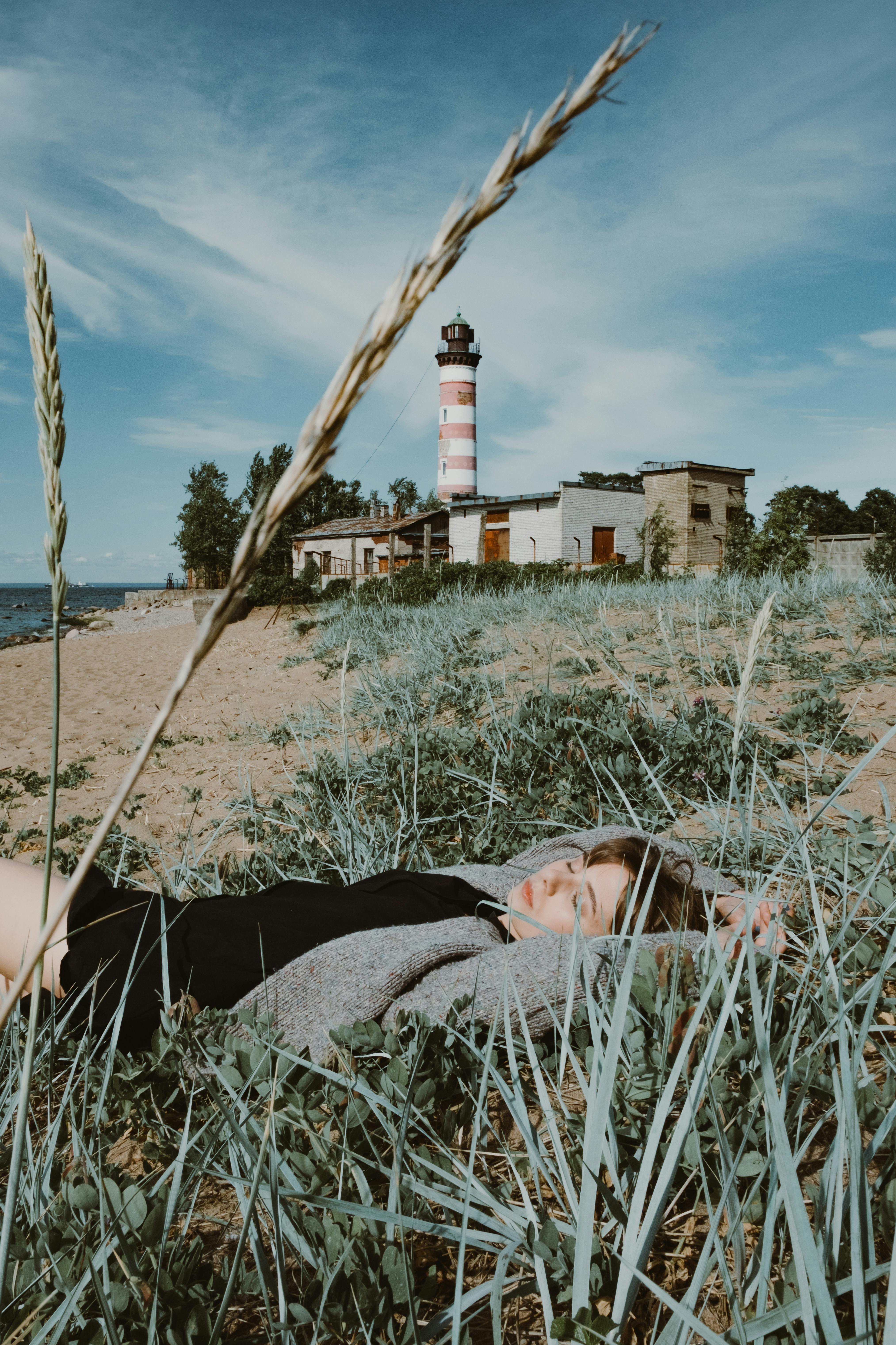 Woman relaxing near a lighthouse on a sunny coastal day, surrounded by grass and sand.