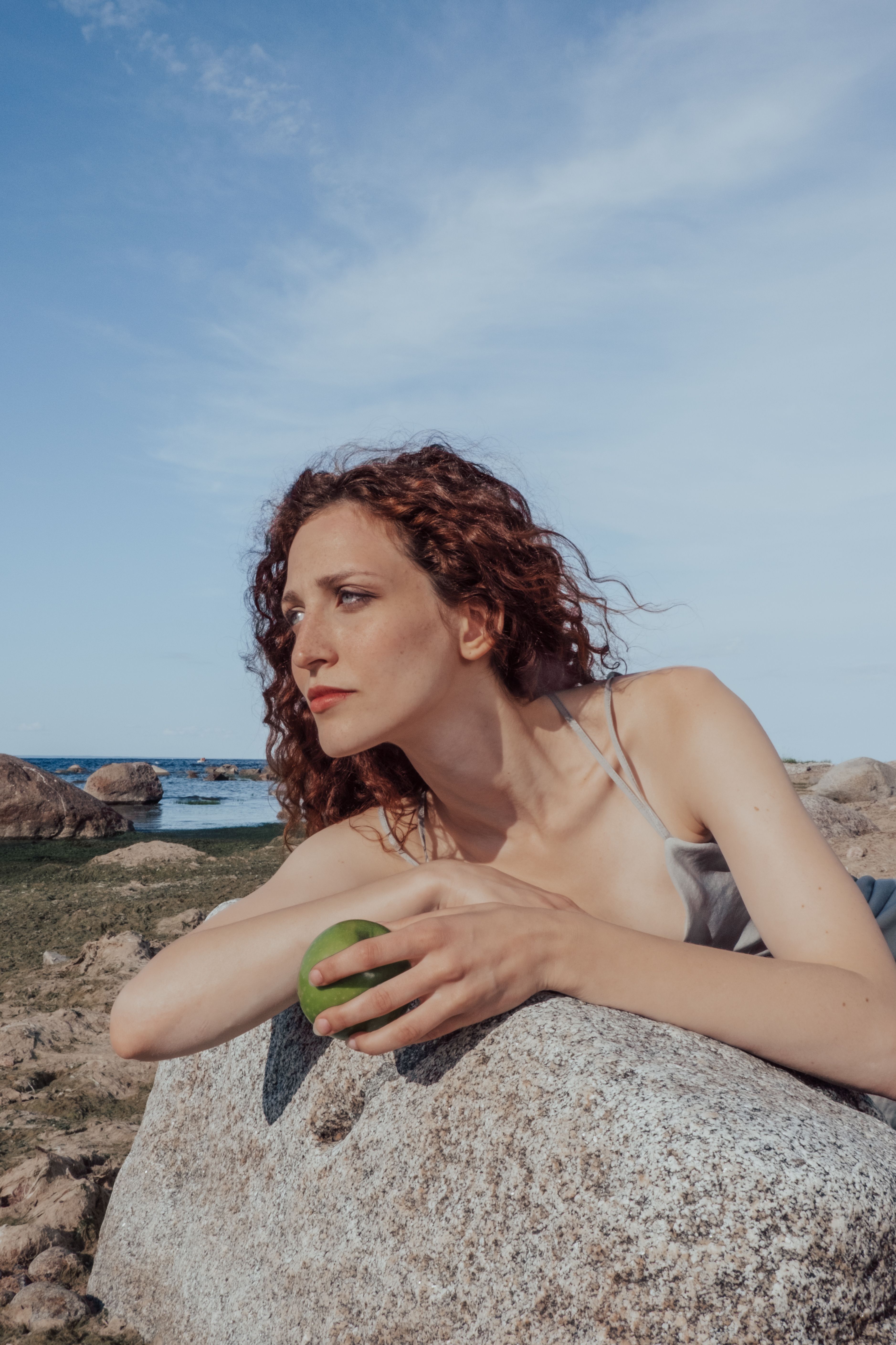 A woman with curly hair holds a green apple while sitting on rocks outdoors under a clear sky.