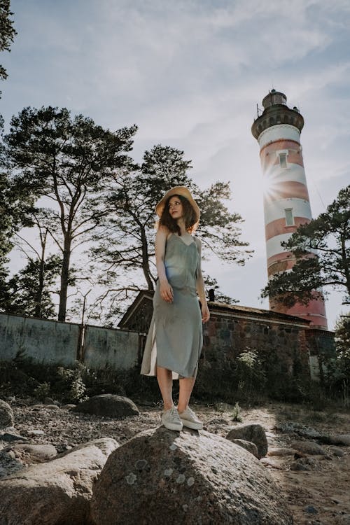 Free Woman in Gray Dress Standing on Rock Near Lighthouse Stock Photo