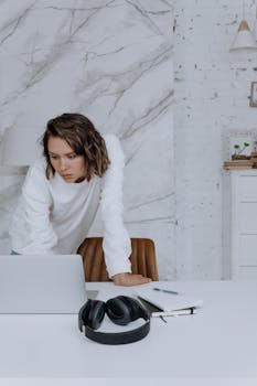 A woman in a white sweater focuses on work at her laptop in a stylish home office setting.