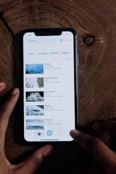 Close-up of hands using a smartphone displaying search results for ocean videos, on a wooden table.