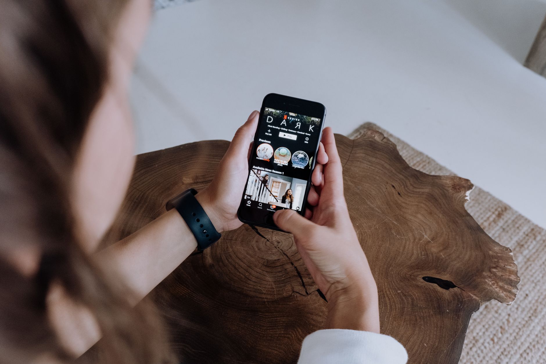 An adult browsing a streaming app on a smartphone while sitting at a wooden table.