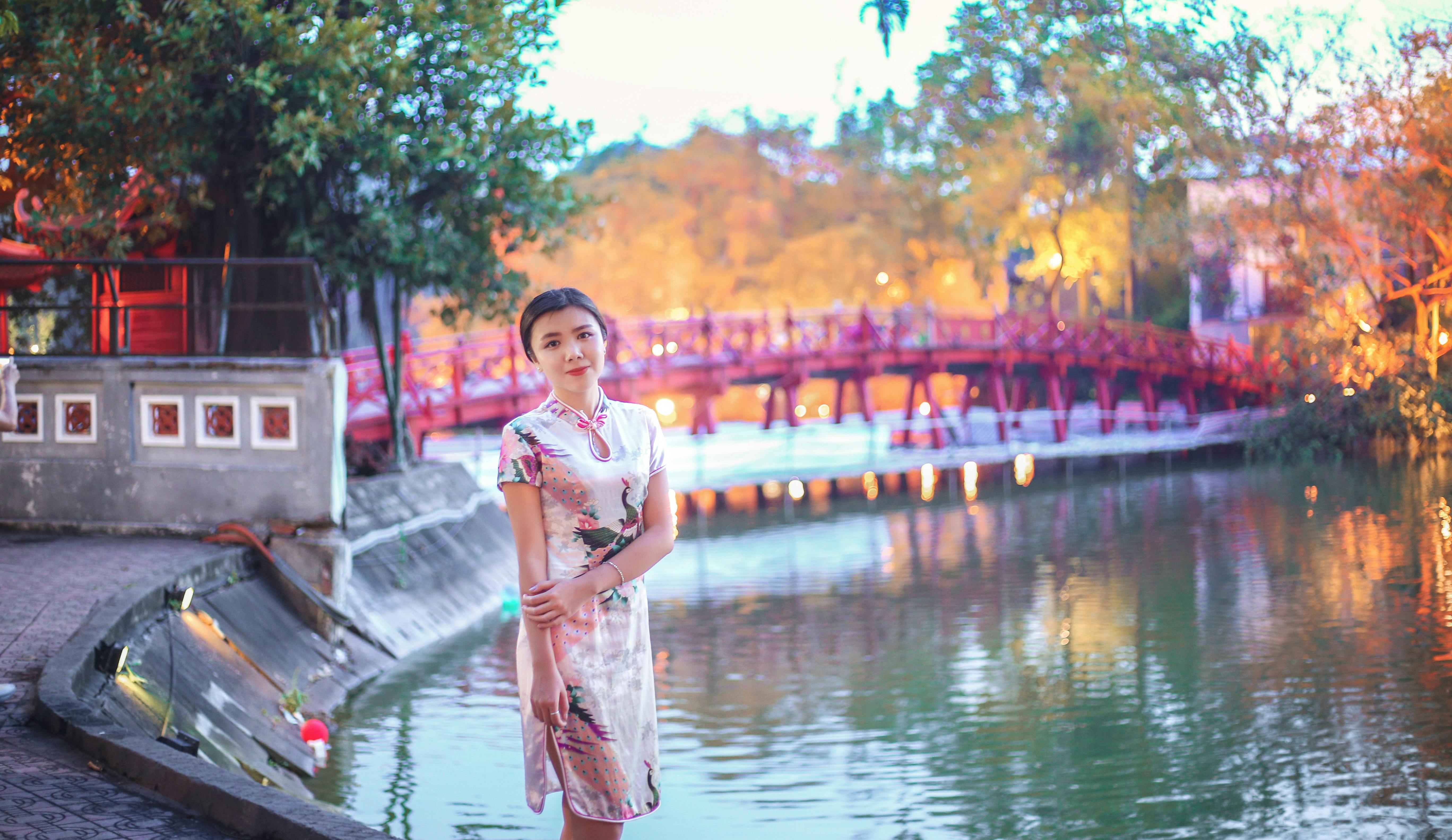 黄昏时分，一位身着传统服饰的女子优雅地立于河边，身后是一座红桥