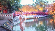 黄昏时分，一位身着传统服饰的女子优雅地立于河边，身后是一座红桥