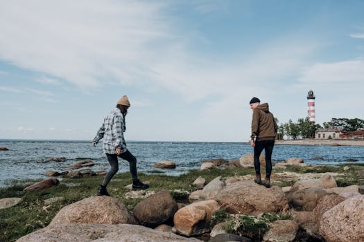 A couple walking on rocks by the sea with a lighthouse in the background.
