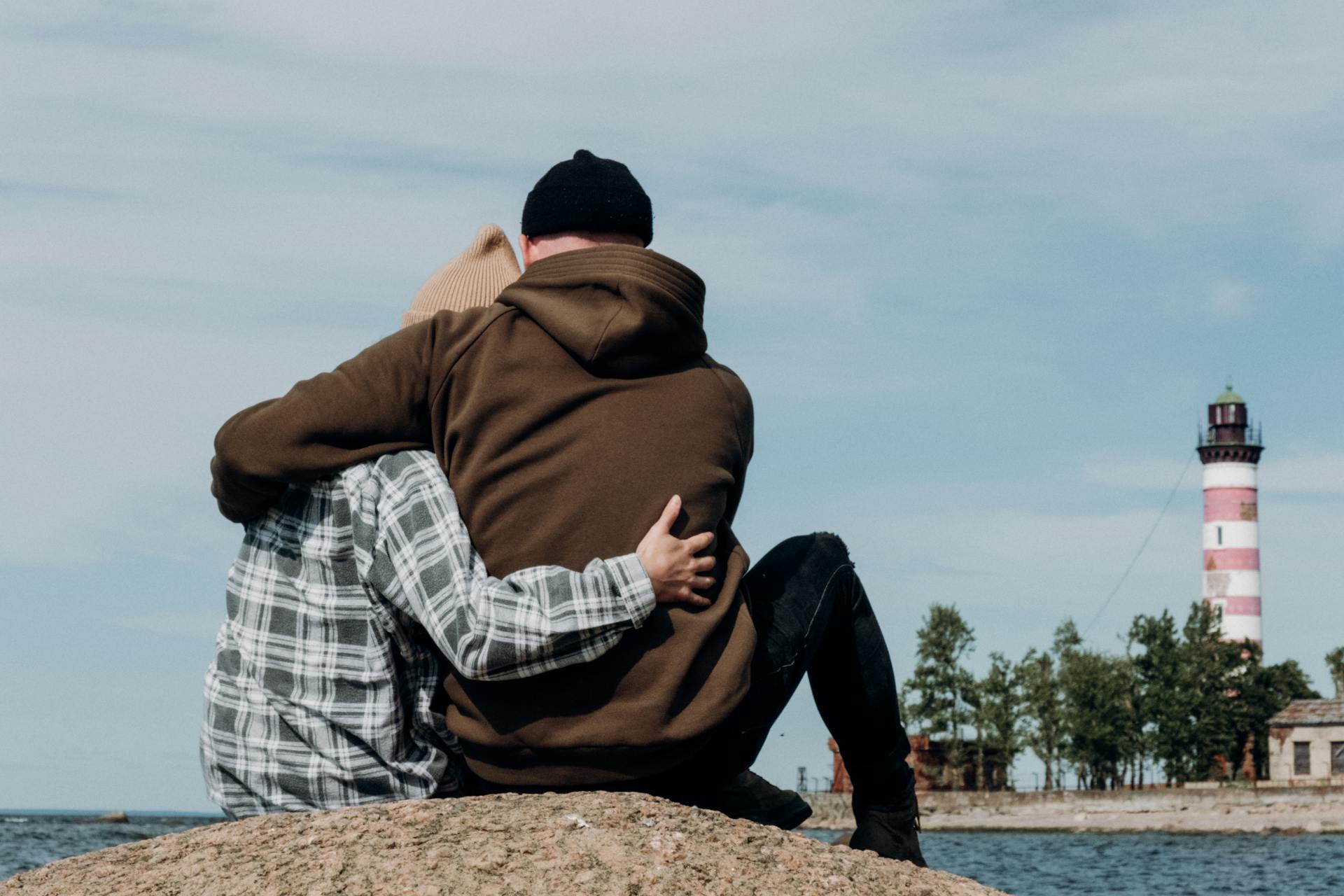 Couple sitting on rocky shore hugging with lighthouse view, evoking romance and tranquility.