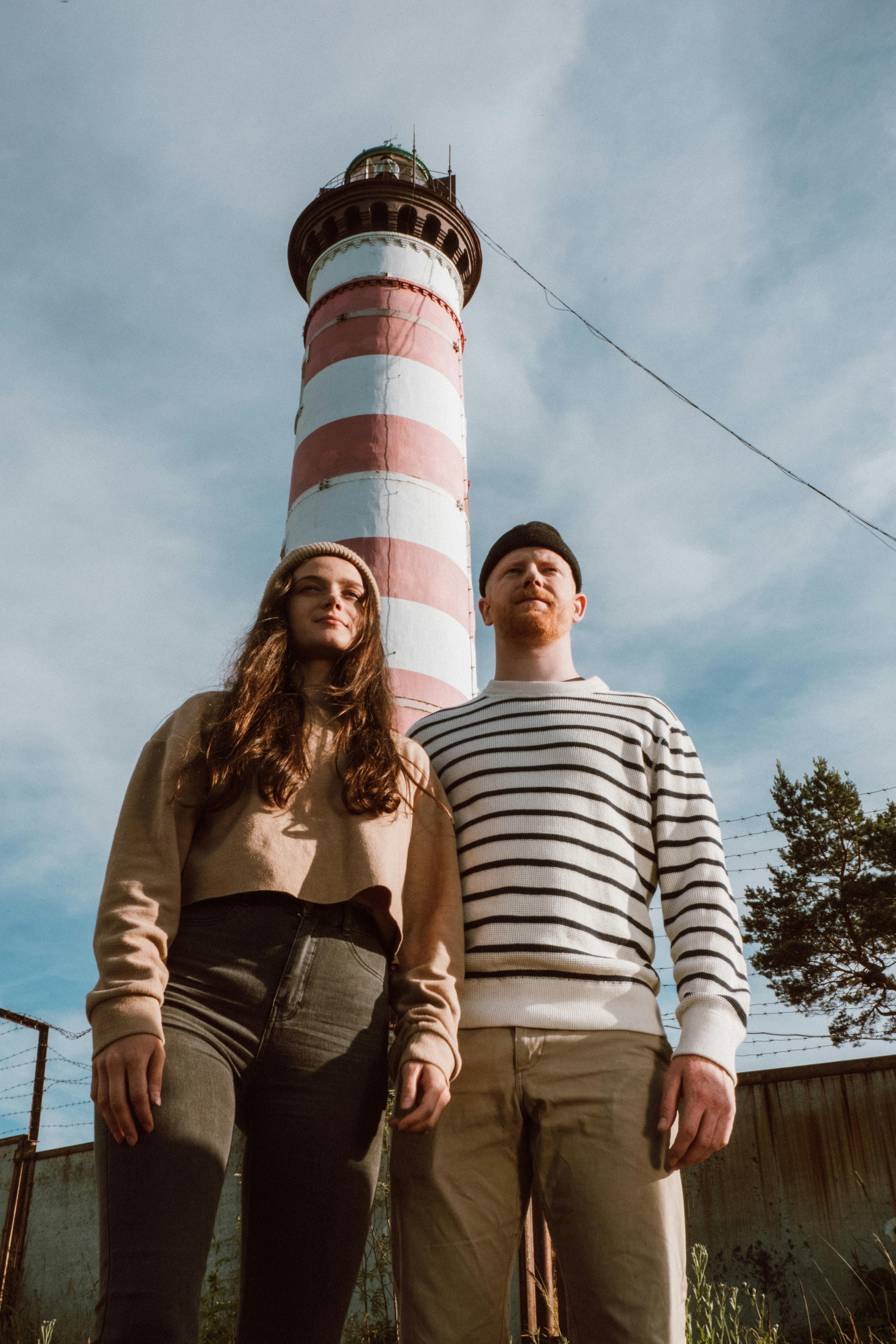 一对年轻夫妇在晴朗的天空下，于高耸的条纹灯塔前摆姿势
