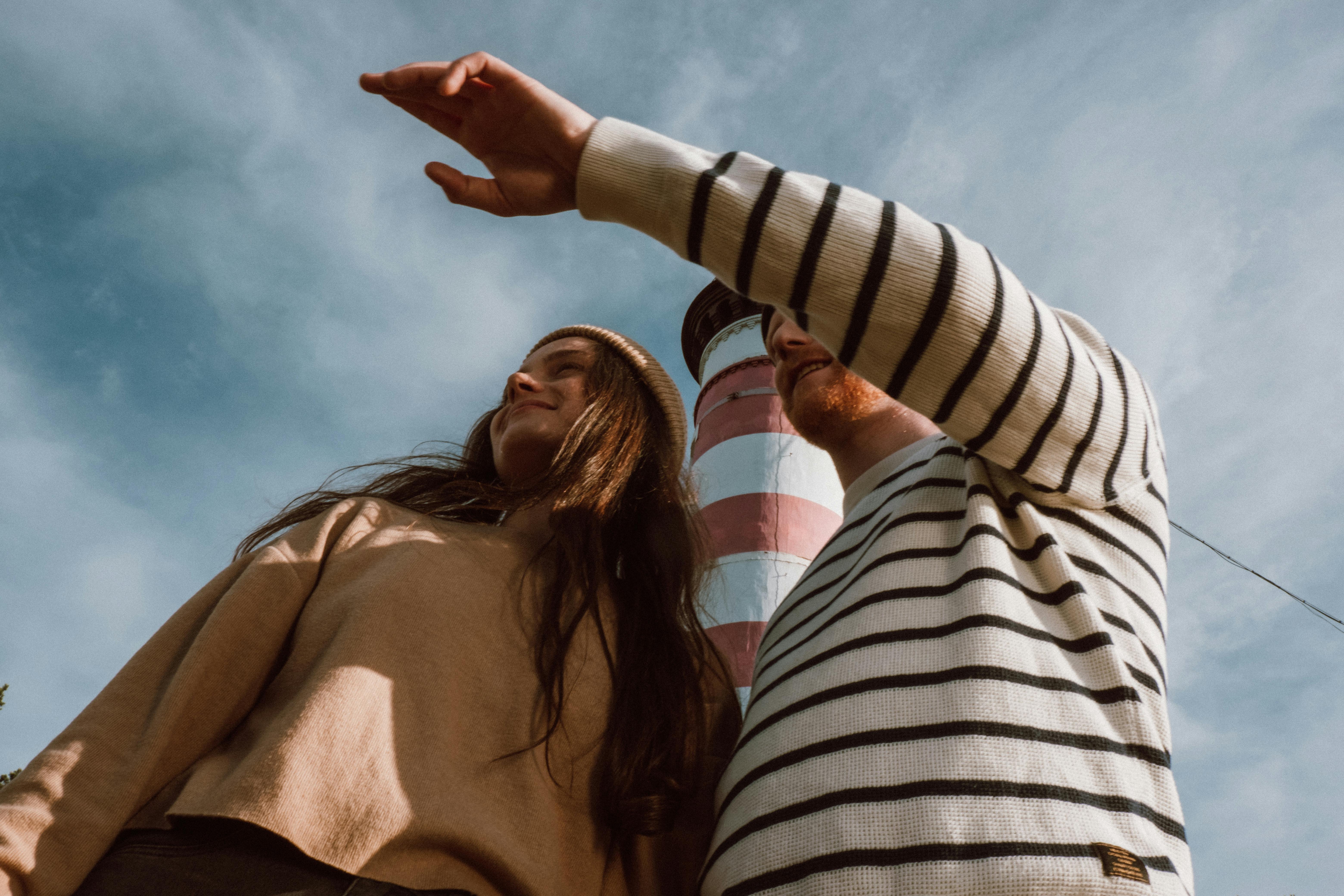 A couple standing near a striped lighthouse, pointing towards the sky with a clear blue backdrop.
