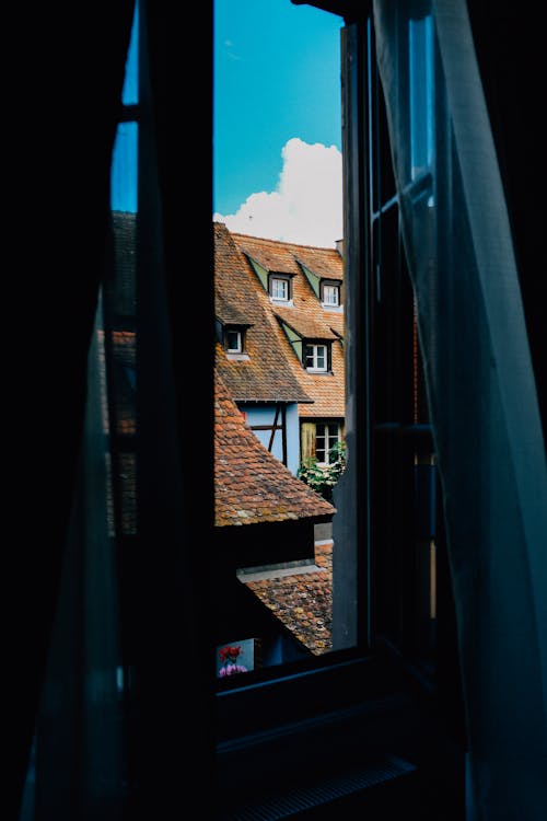Free View through open window with curtains on residential buildings with windows on tiled roofs against cloudy sky in old town Stock Photo