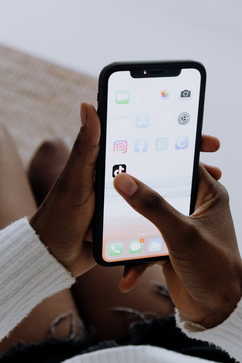 A person holding a smartphone in a dim room, thumb hovering over a TikTok feed showing colorful short videos with bold on-screen text captions
