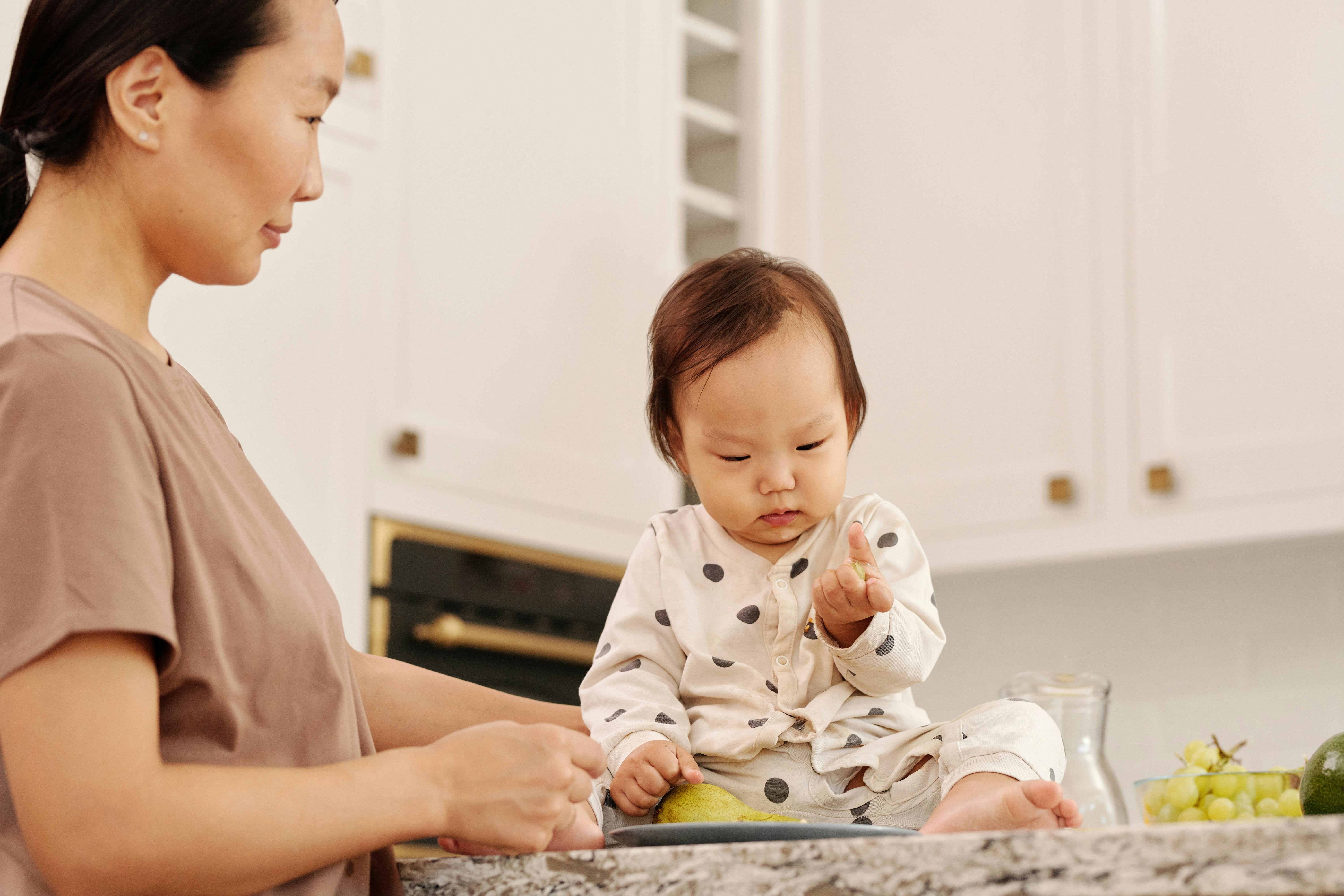在明亮而温暖的厨房里，一位亚洲母亲正与女婴分享着温柔的时刻