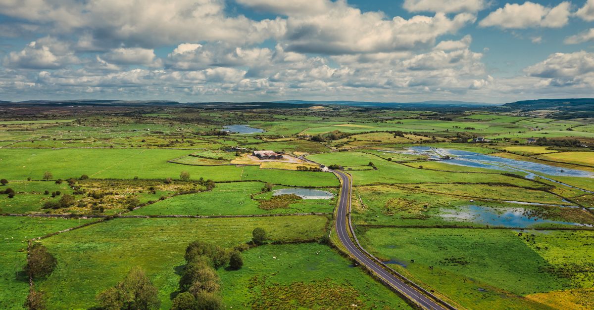 Aerial view of the vibrant green countryside and long road in County Clare, Ireland, under a partly cloudy sky.