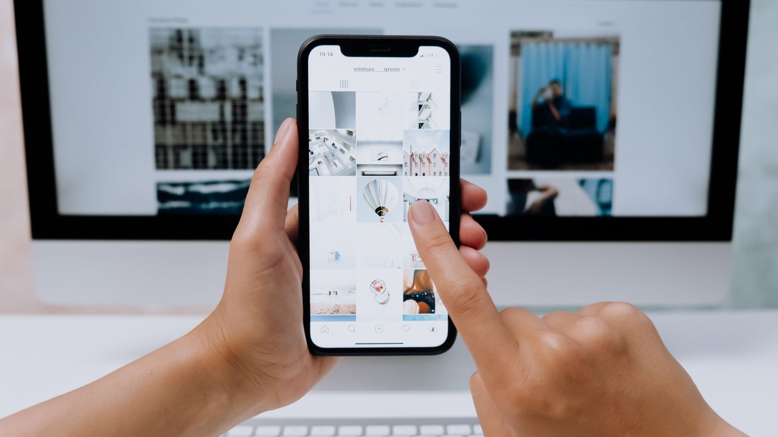 Hands scrolling a photo grid on a smartphone in front of a computer
