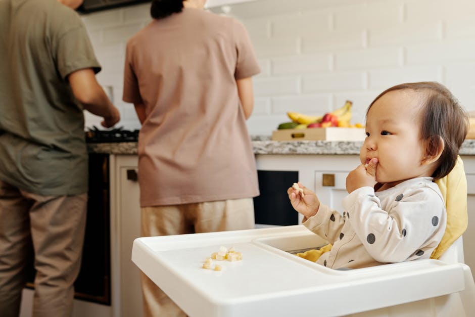 Cute toddler sits in a high chair enjoying a meal, while parents cook in the background of a modern kitchen.