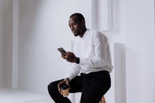 Free Businessman in a white shirt uses a smartphone indoors with a coffee cup. Stock Photo