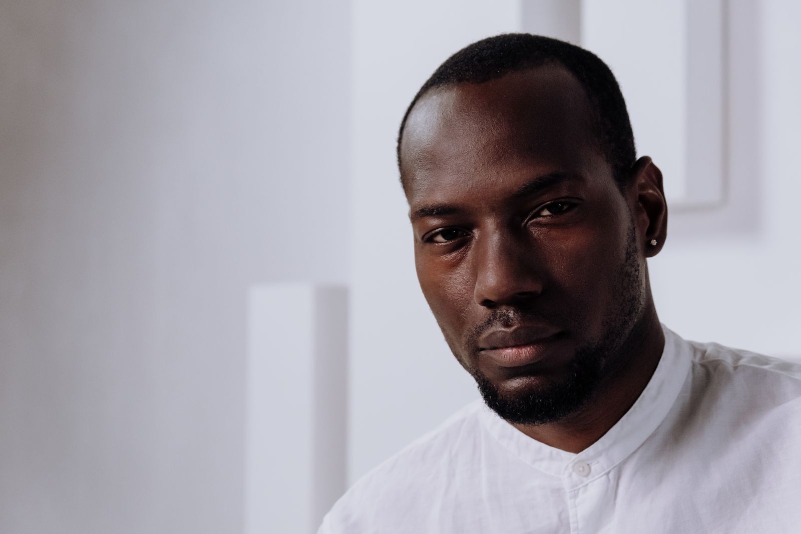 Close-up studio portrait of a Black man in a white shirt with a calm, reflective expression, soft natural light against a pale wall.
