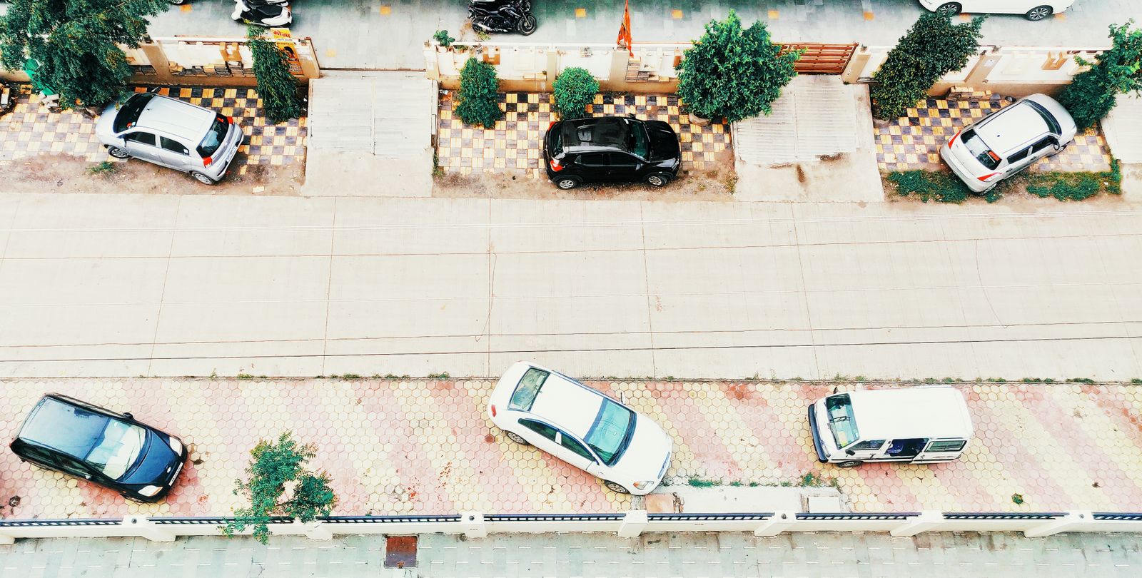 Aerial shot of an urban street in Indore, India, featuring parked cars along a tiled sidewalk and a quiet roadway.