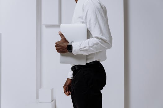 Professional adult holding a laptop in a modern, minimalist office setting with electronic watch.