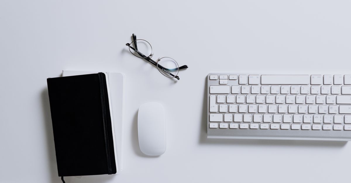A clean and modern workspace featuring a keyboard, mouse, notebook, and eyeglasses arranged on a white desk.