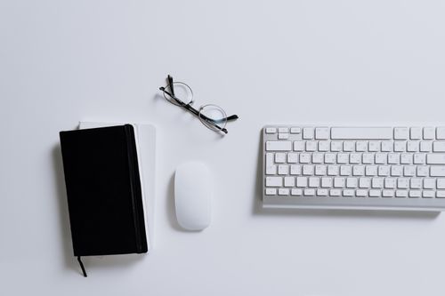 Free A clean and modern workspace featuring a keyboard, mouse, notebook, and eyeglasses arranged on a white desk. Stock Photo