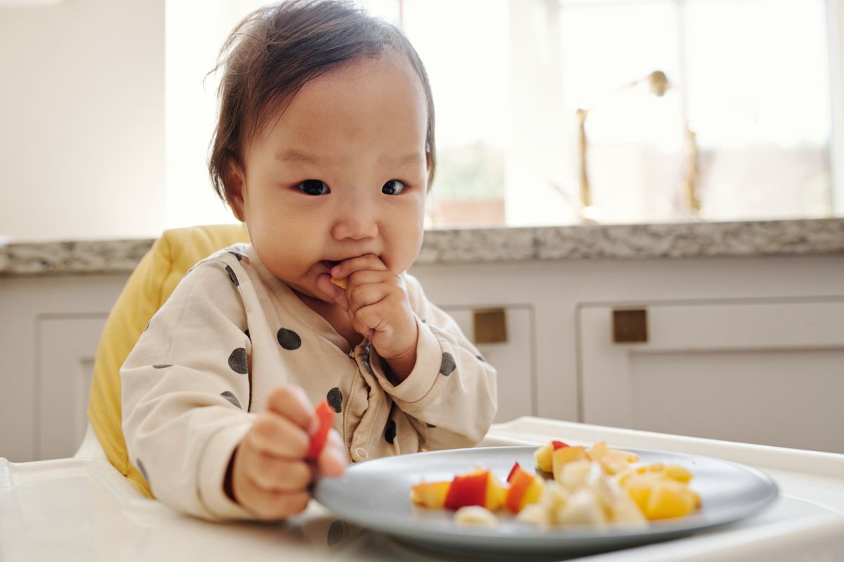 Baby in a polka dot outfit enjoying fresh fruit pieces while sitting in a highchair at home