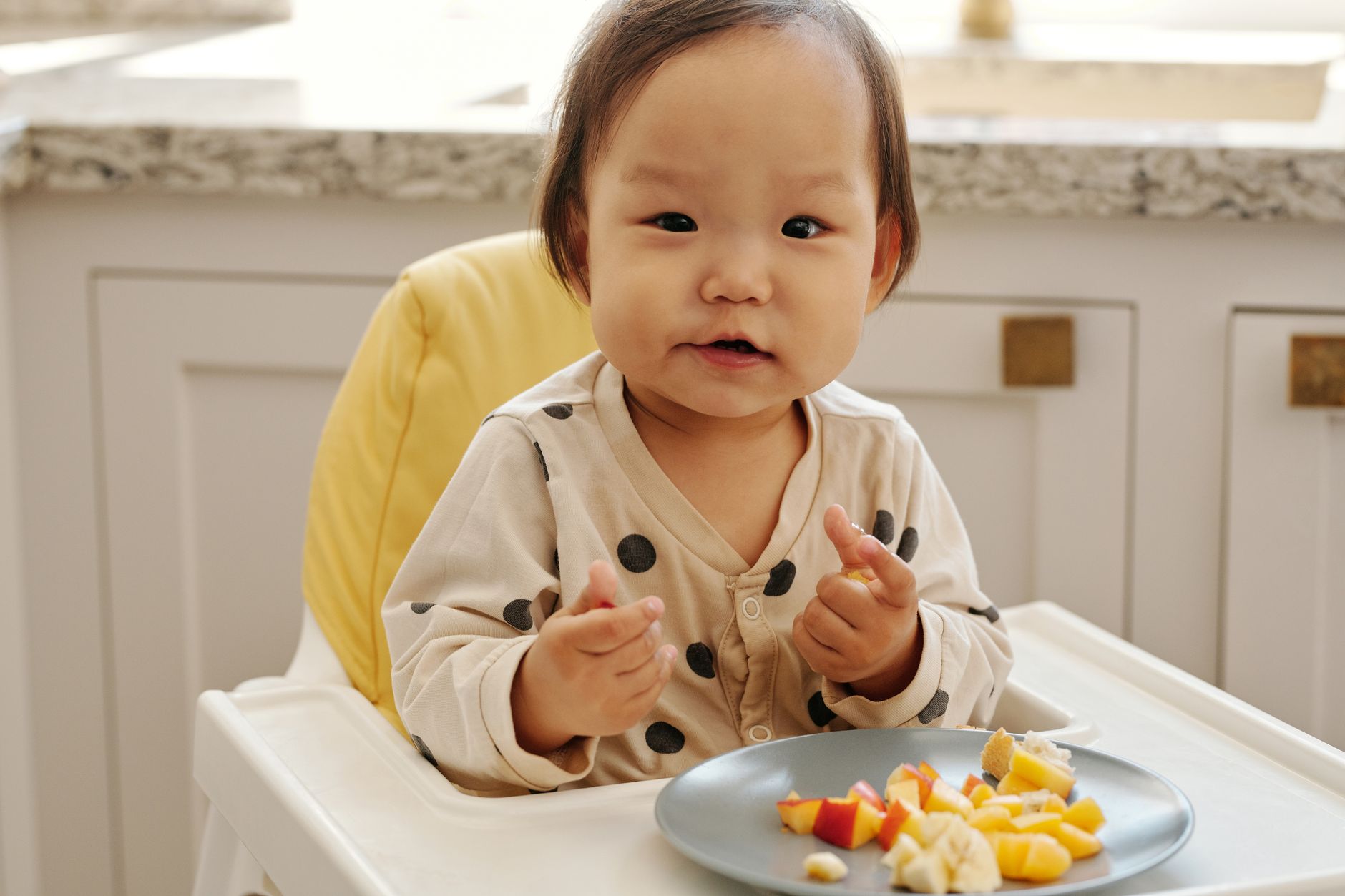 Cute toddler in polka dot outfit enjoys healthy fruit snack indoors, sitting in high chair.