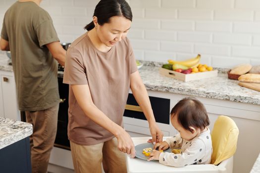 母亲在现代化的厨房里，给坐在高脚椅上的幼儿喂健康水果