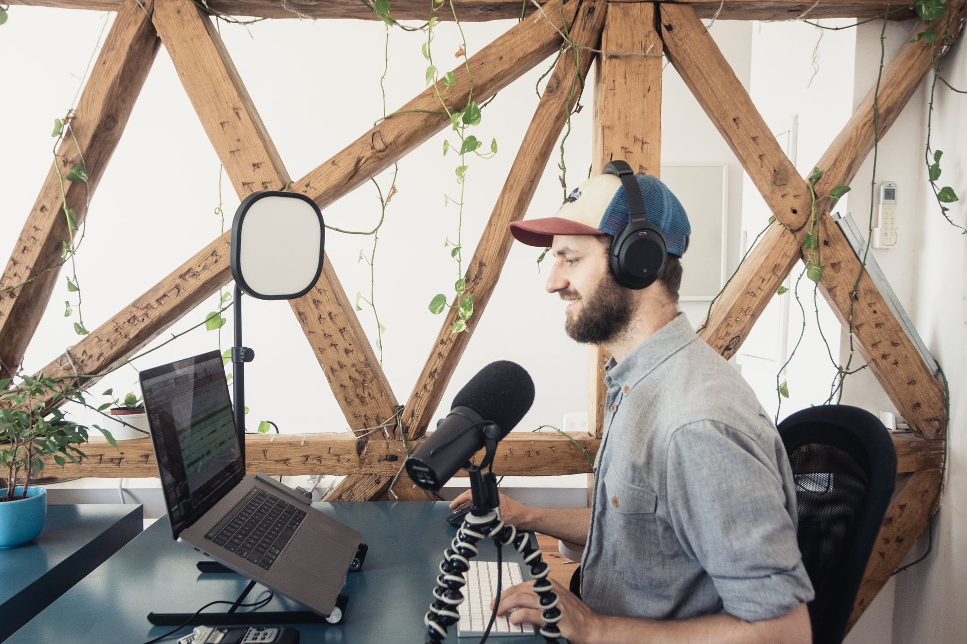 A man with headphones podcasts at a desk with a laptop and microphone stand indoors.