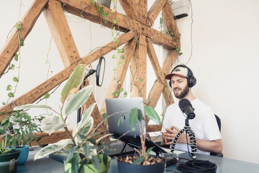 A man in a white shirt records a podcast with a laptop and microphone in an indoor setting with plants.
