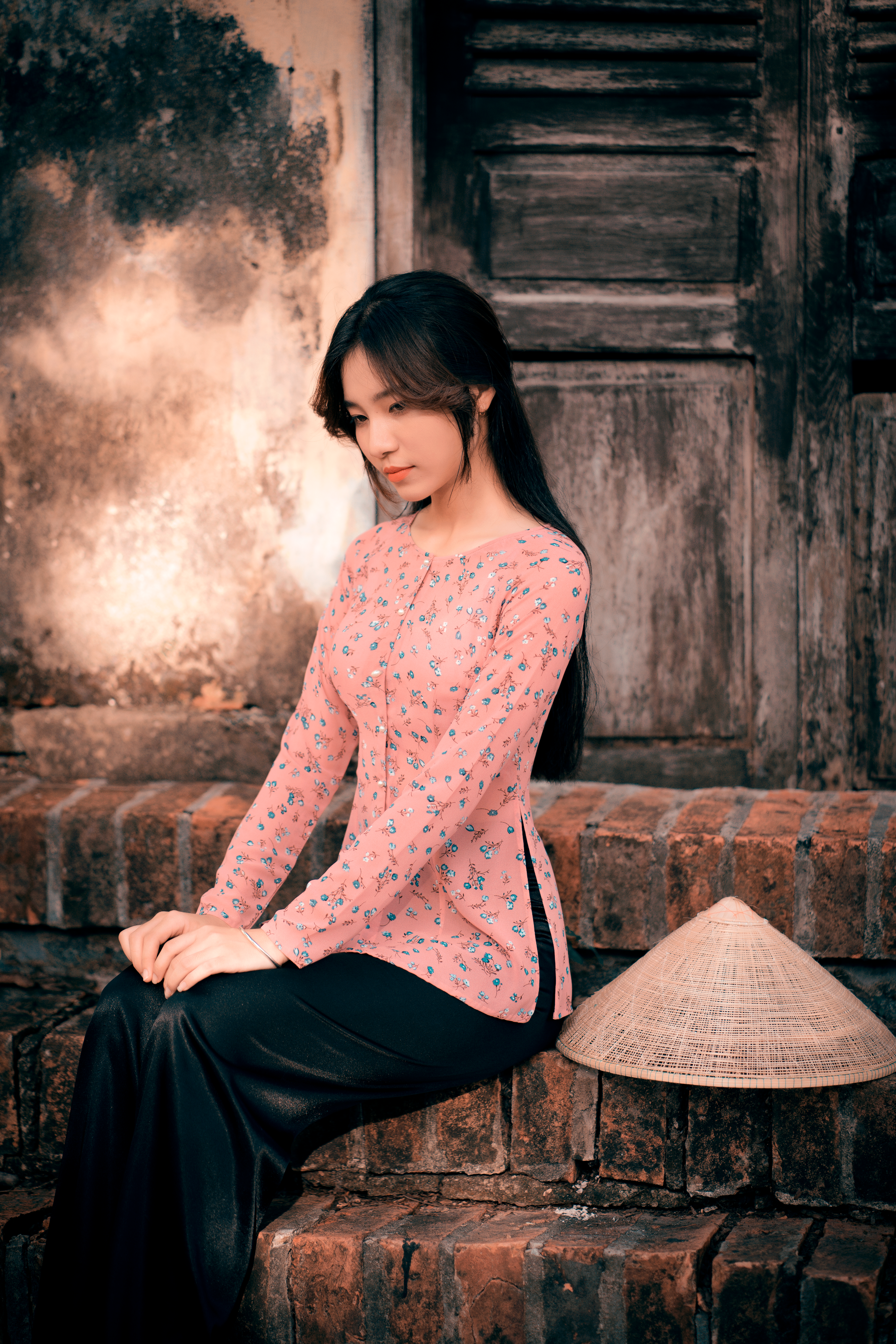 一名身穿花卉衬衫的年轻女子，头戴圆锥帽，坐在户外的砖块上，散发着宁静优雅的气质