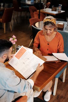 A couple reviews menus while dining indoors at a cozy restaurant, engaging in conversation.