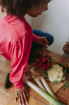 A woman in a red blouse sits at a wooden table arranging fresh vegetables including radishes and leeks.