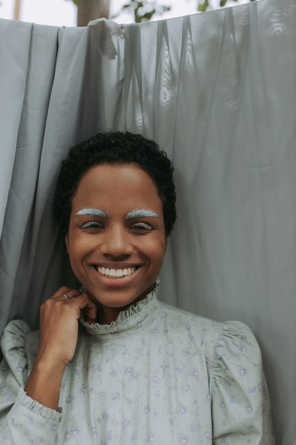 A joyful woman with natural hair and unique makeup smiles warmly while standing behind a curtain.
