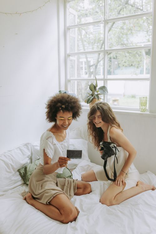 Free Two women smile as they sit on a bed, sharing instant photos in a bright bedroom. Stock Photo