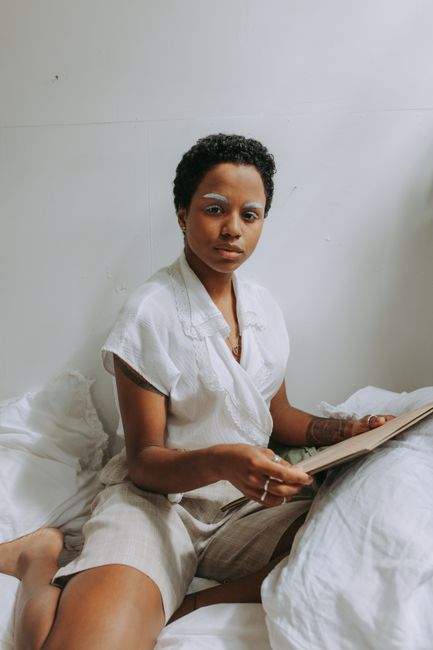 A thoughtful young woman sits on a bed, holding a folder, looking directly at the camera.