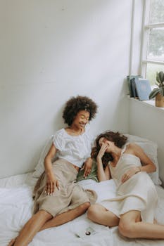 两个女人在明亮舒适的卧室里，躺在床上笑着放松，自然光线充足