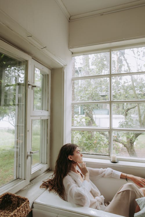 Woman in White Long Sleeve Shirt Sitting on Bed Near Window