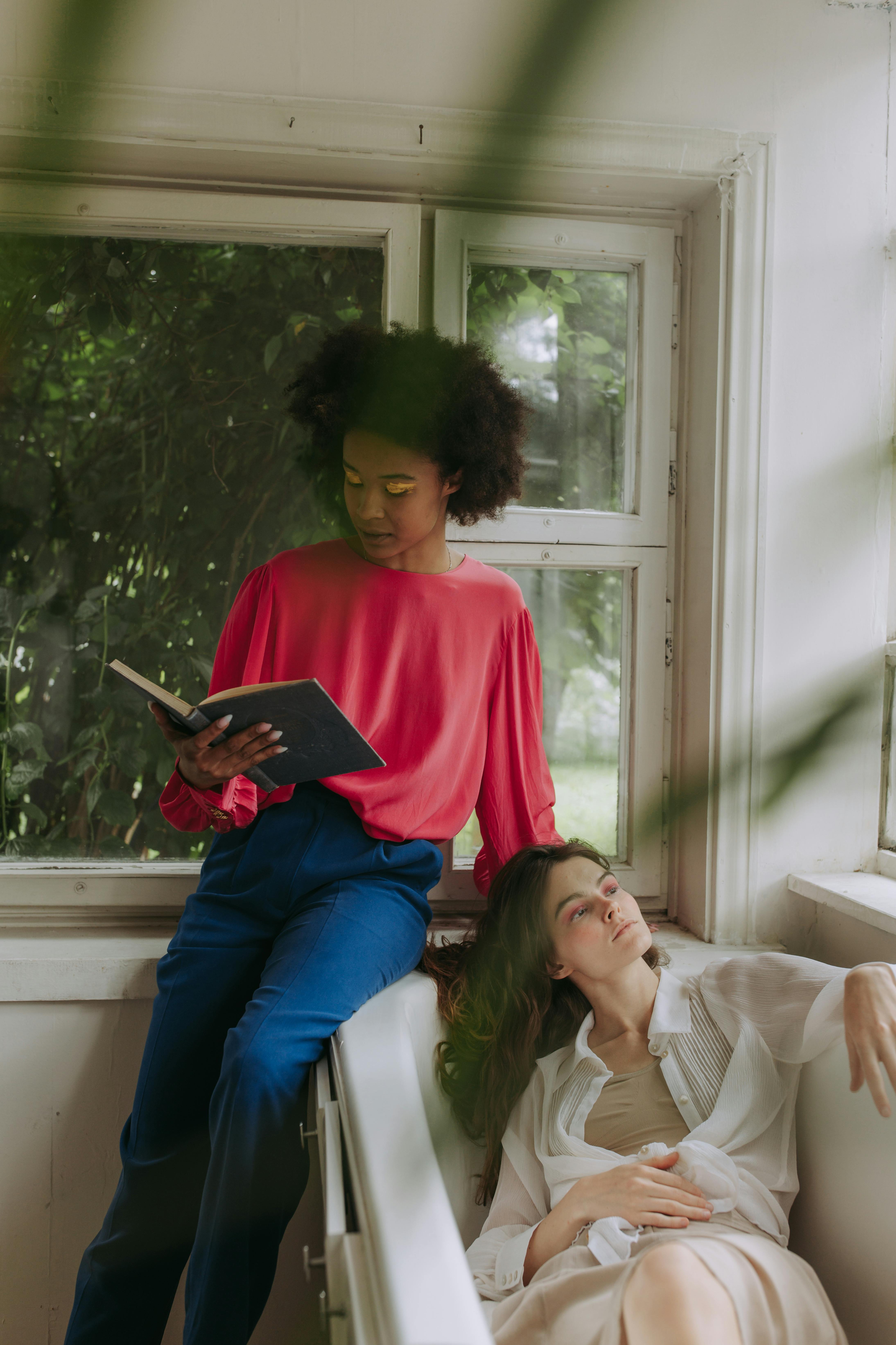Two women enjoying a tranquil moment with a book by the window in a bright indoor space.